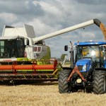 A modern combine harvester and tractor working together to harvest wheat in a sunny outdoor field.