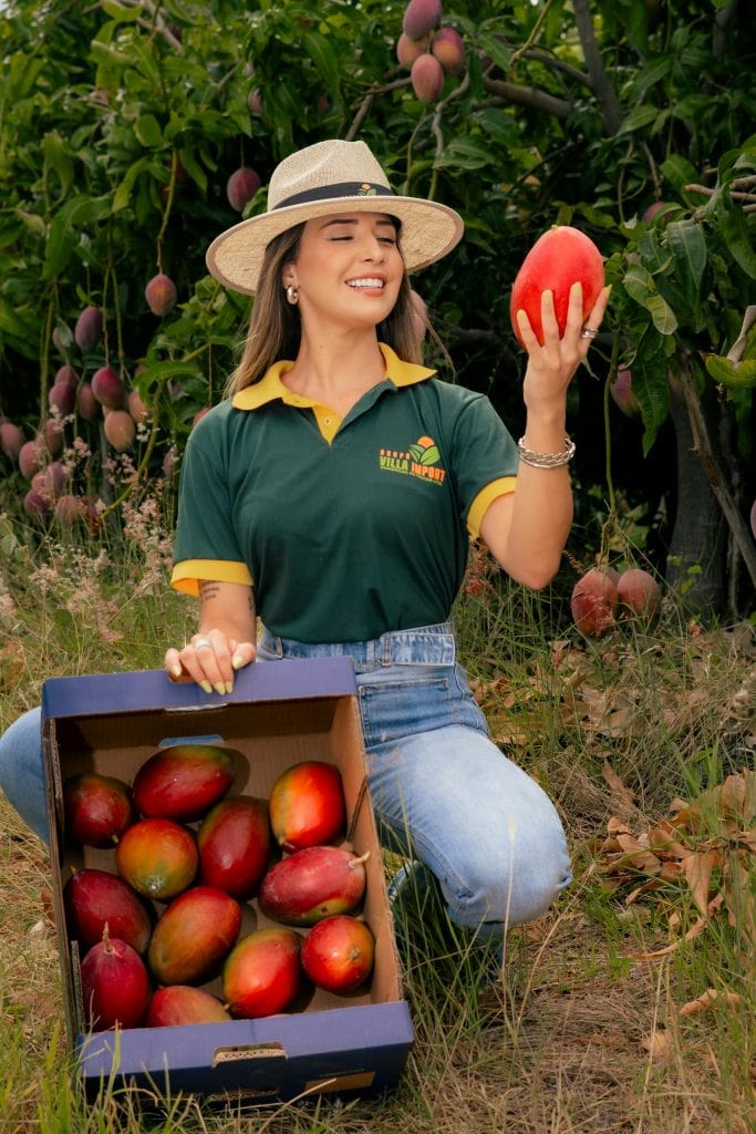 Smiling woman in hat and green shirt holding fresh mango in orchard with box of fruits.
